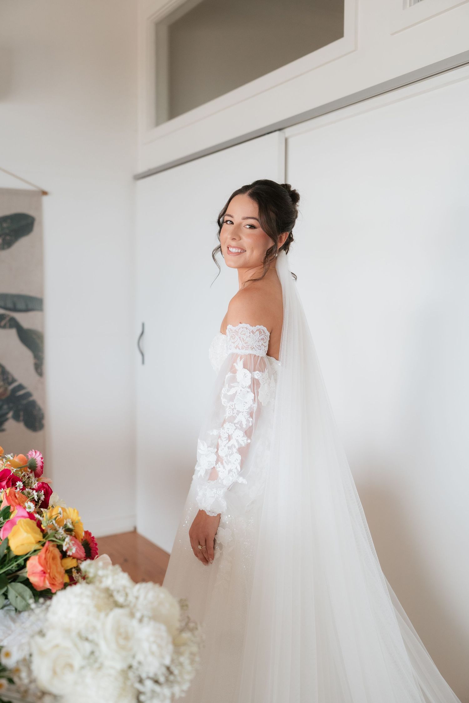 Bride in off-shoulder lace wedding gown with veil smiles indoors near colorful floral bouquet.