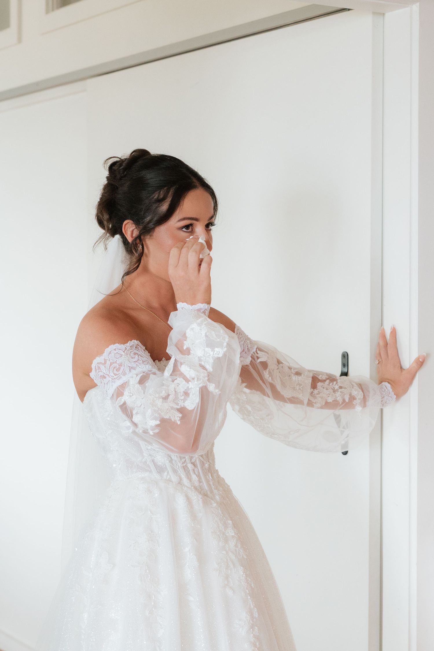 Emotional bride in off-shoulder lace wedding gown with sheer sleeves wiping tears near white door.