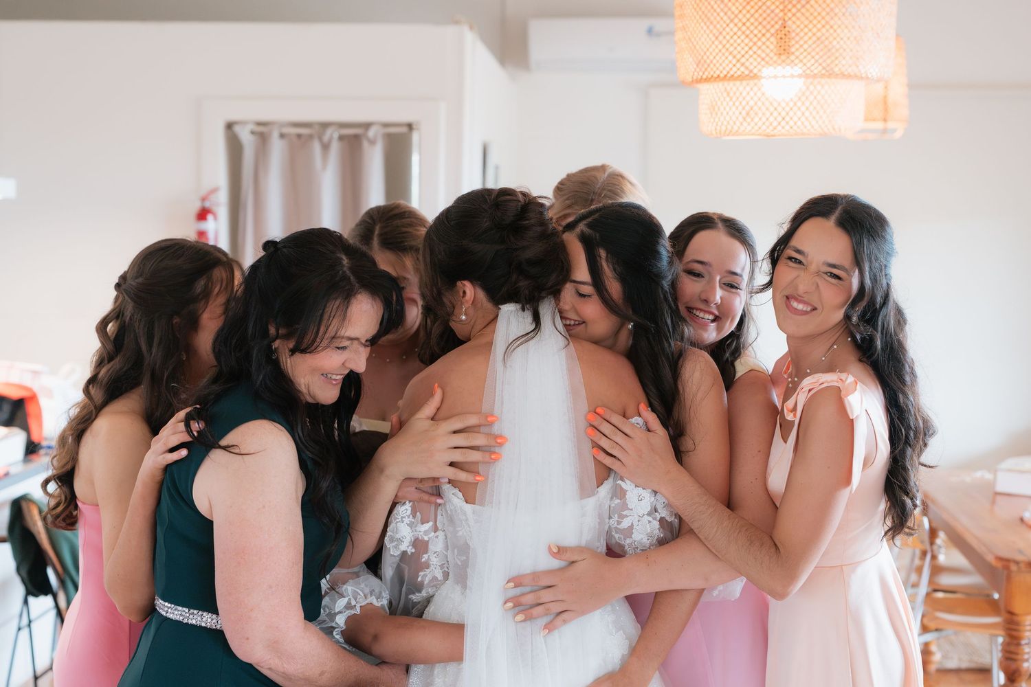 Bridesmaids in pink and green dresses share a heartfelt group hug with the bride in her white lace wedding gown.