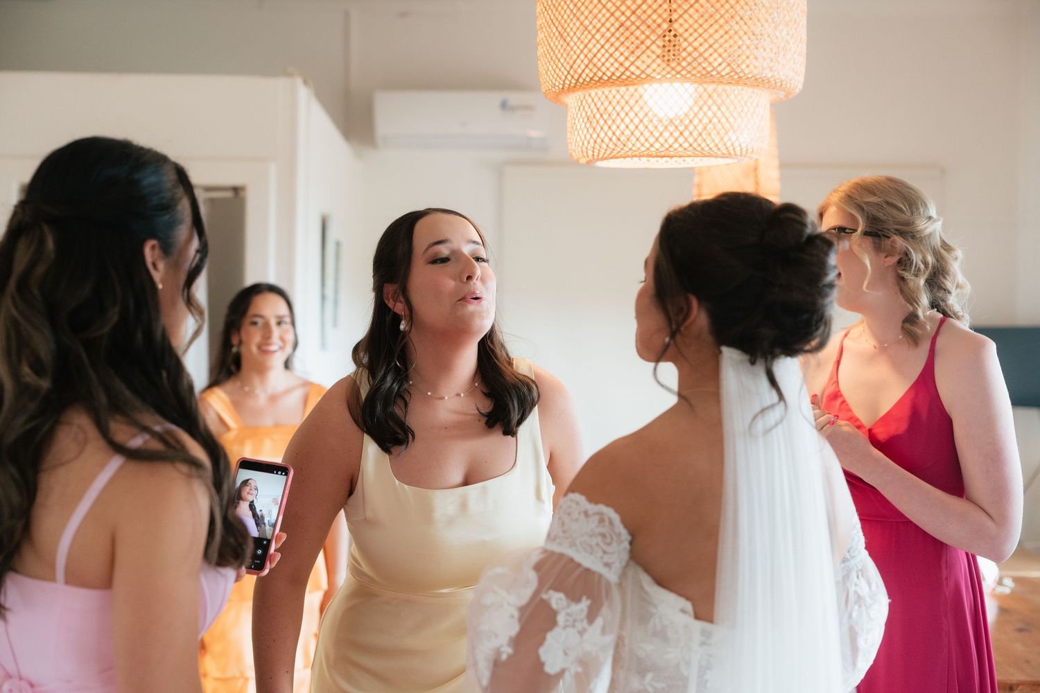 Bridesmaids in colorful dresses surround a bride in a white gown during wedding preparations indoors.