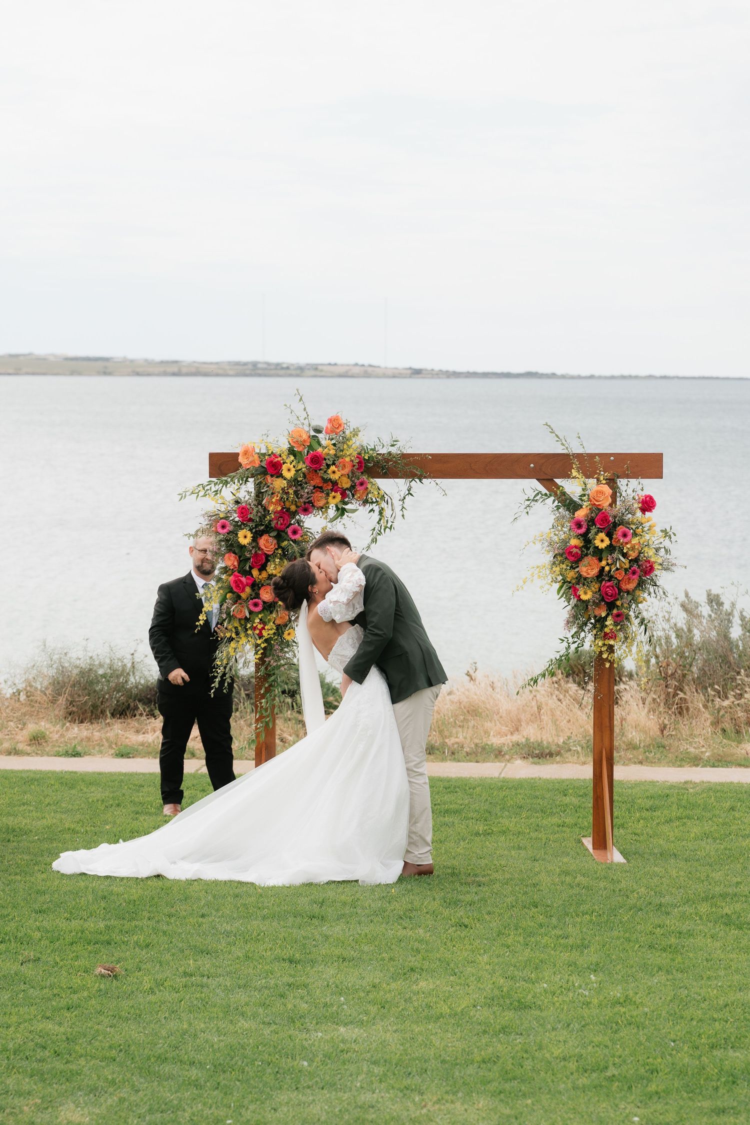 Bride and groom share first kiss under a wooden arch with colorful floral arrangements by the waterfront.
