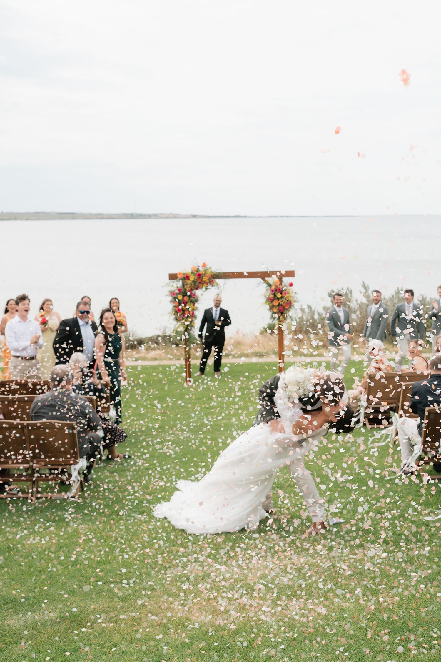Couple shares romantic kiss during outdoor waterfront wedding ceremony with confetti and floral arch backdrop.