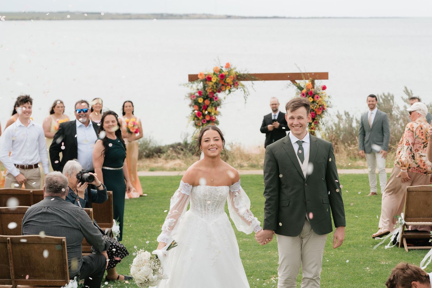 Bride and groom walk down the aisle at outdoor waterfront wedding ceremony with floral arch backdrop.