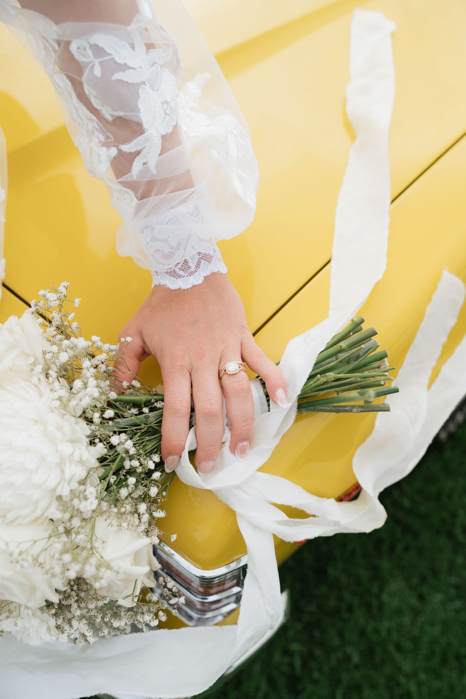 Bride in lace wedding dress holding white rose bouquet with ribbon on yellow vintage car hood.