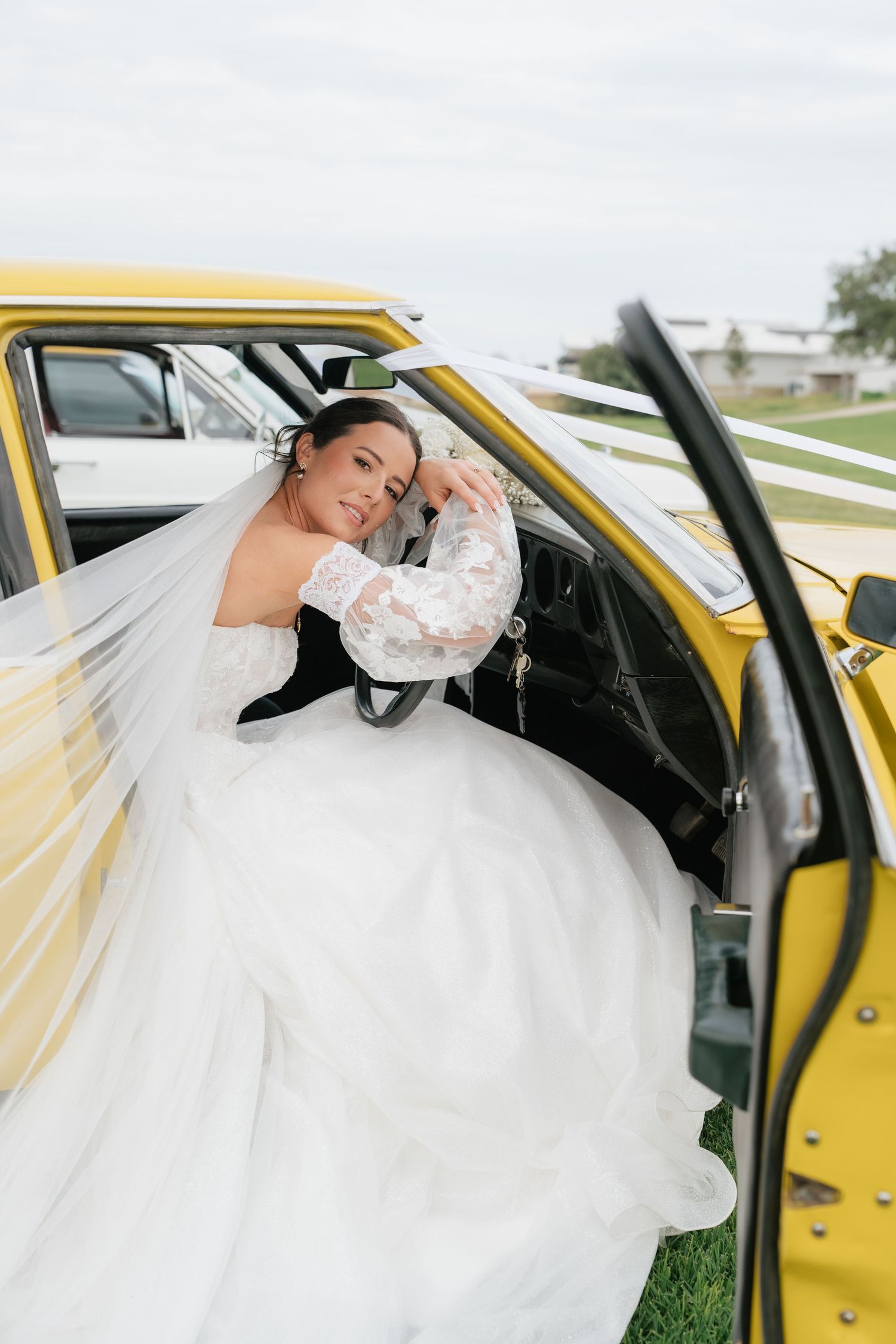 Bride in white lace gown and veil poses inside a yellow vintage truck door on a green lawn.