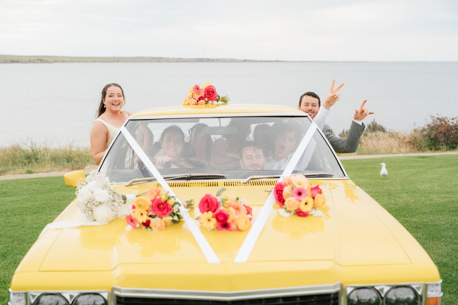 Bride and groom celebrating in a vintage yellow car decorated with colorful floral bouquets by the waterfront.