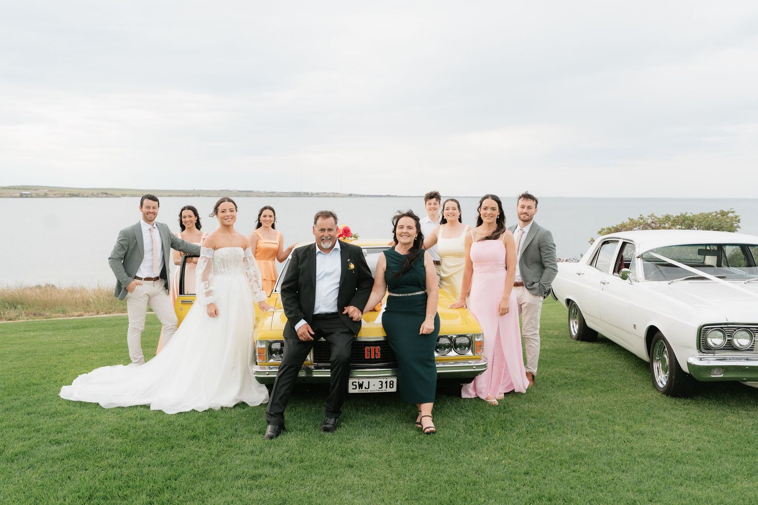 Wedding party poses with vintage yellow car near waterfront, bride in white gown, bridesmaids in pink and green dresses.