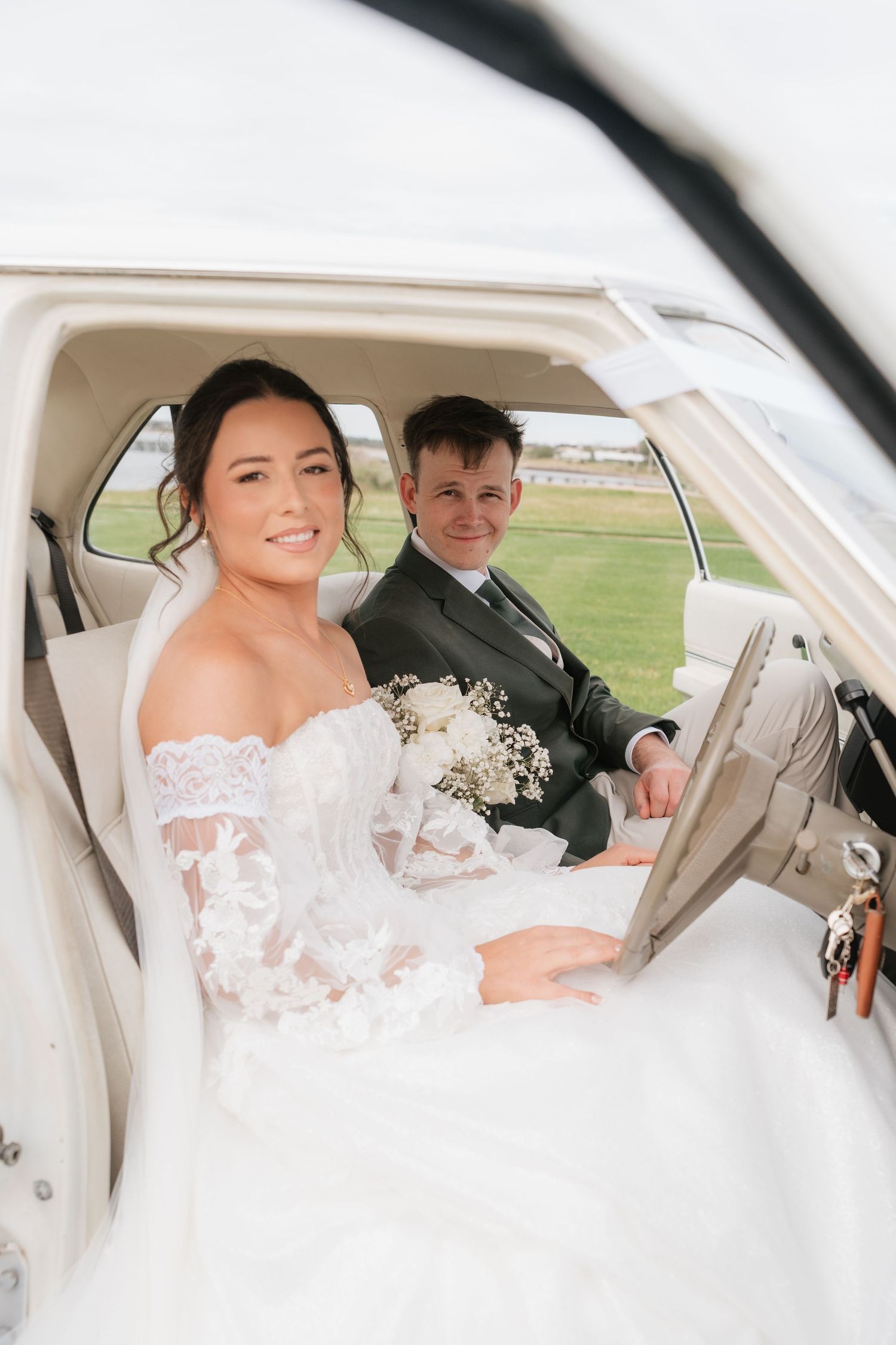 Bride and groom smiling inside vintage white car on their wedding day with bouquet.