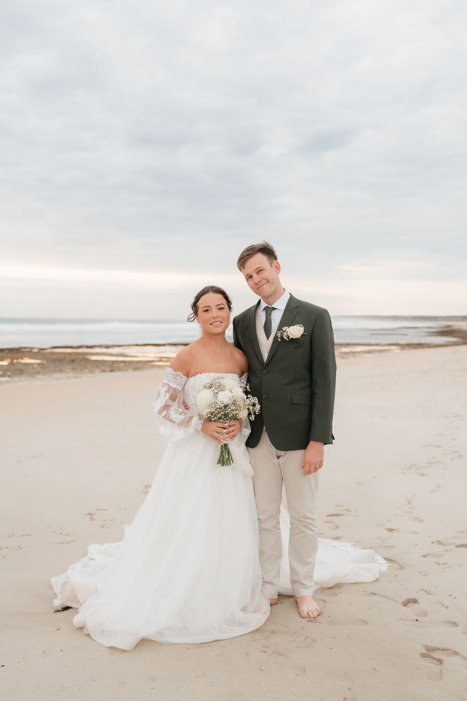 Bride and groom posing together on a sandy beach, bride holding a white bouquet in an off-shoulder lace gown.