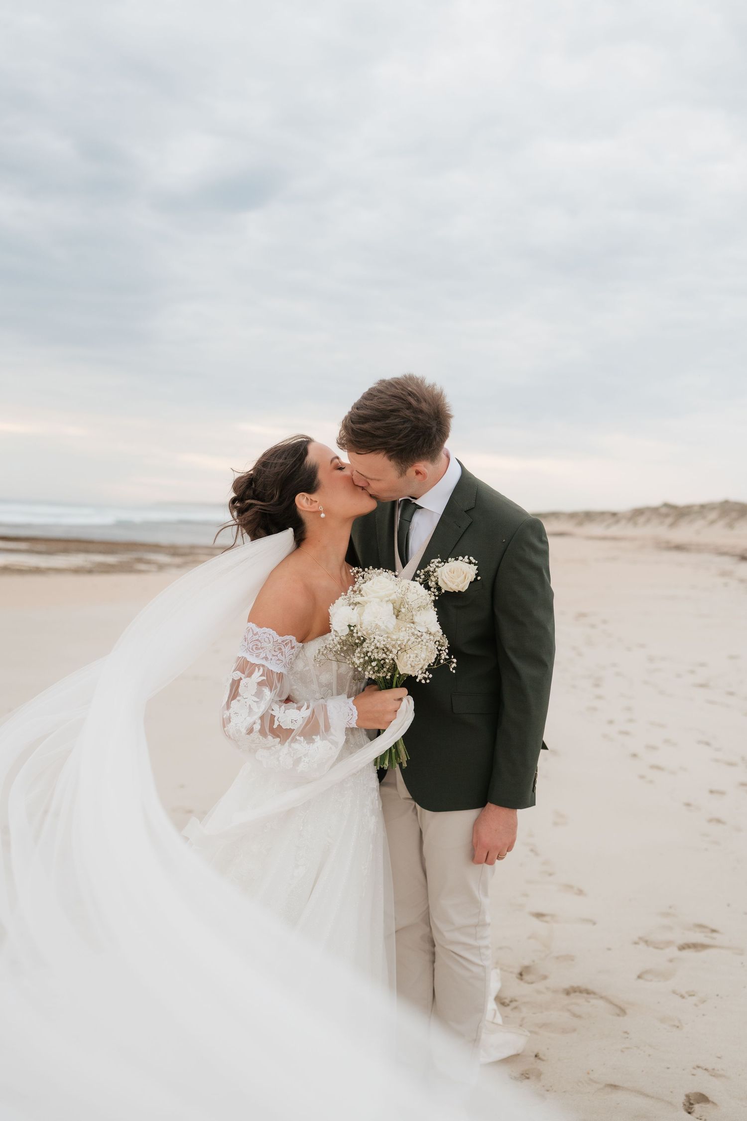 Bride and groom share a romantic beach kiss, her veil flowing in the wind, holding a white bouquet under a cloudy sky.
