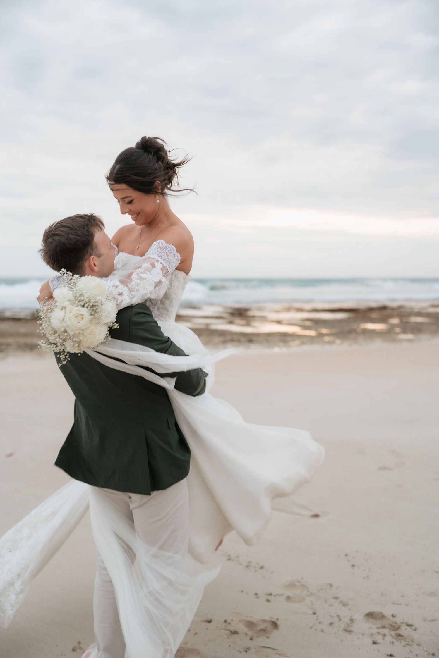 Groom lifting bride in white lace dress on sandy beach with ocean waves in background.