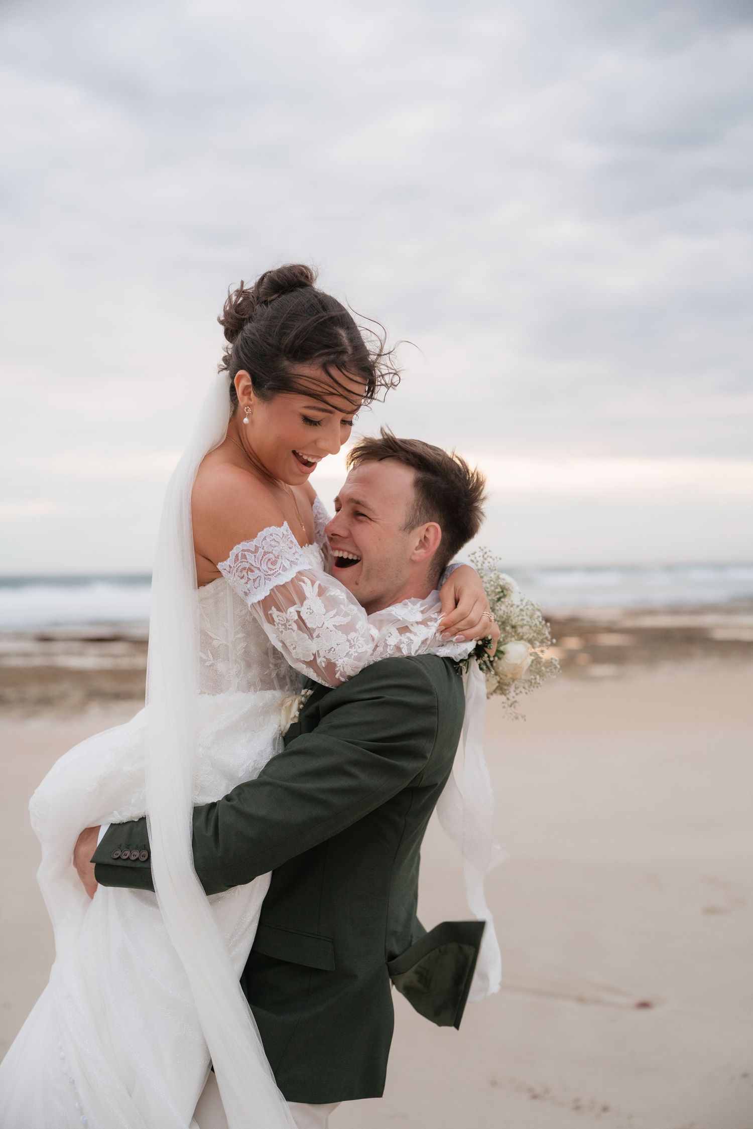 Groom lifting bride on beach, both laughing joyfully, bride in lace gown, groom in green suit.