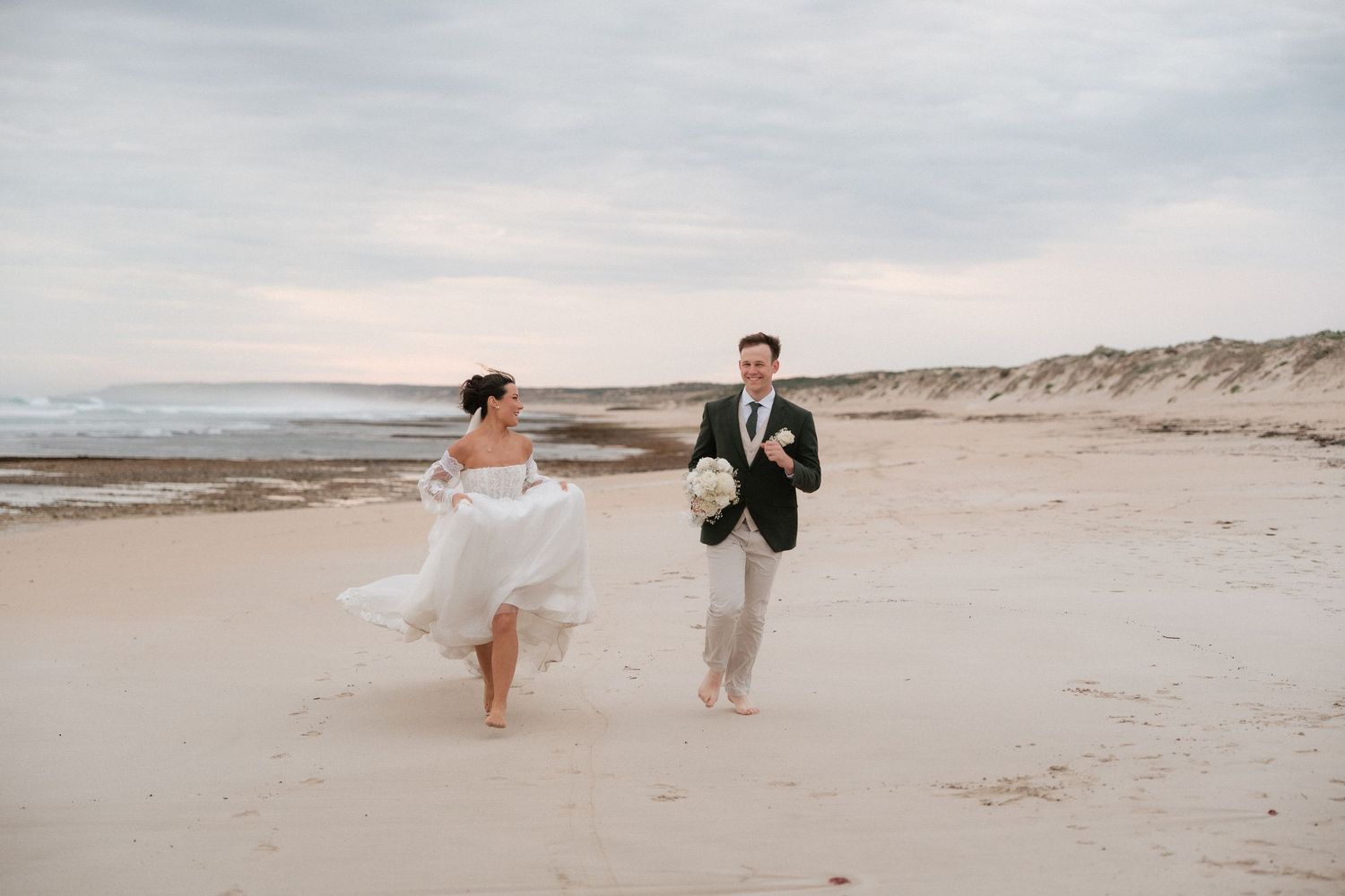 Bride and groom walk barefoot on sandy beach with ocean waves and dunes in background.