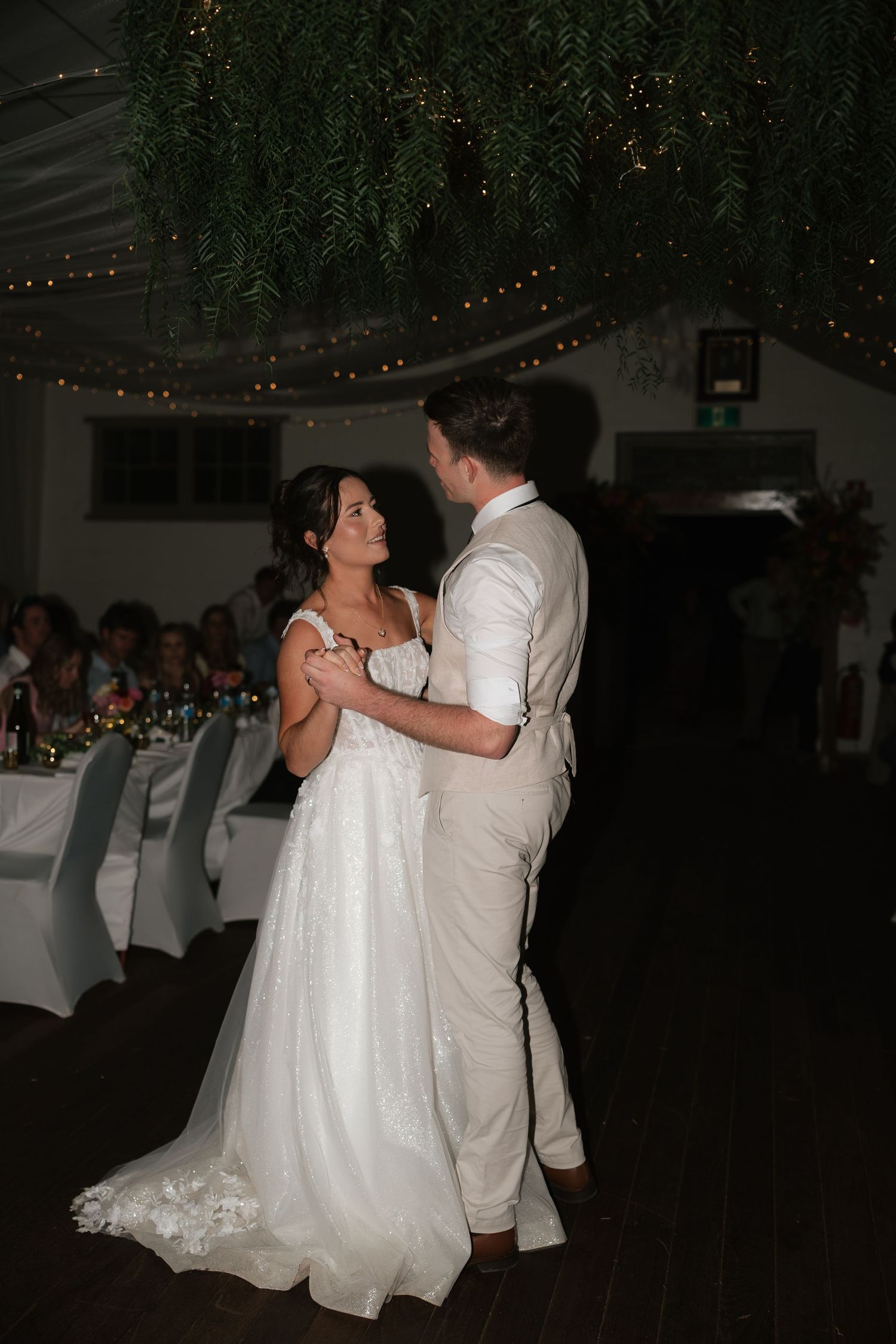 Bride and groom share romantic first dance under fairy lights and greenery at wedding reception.