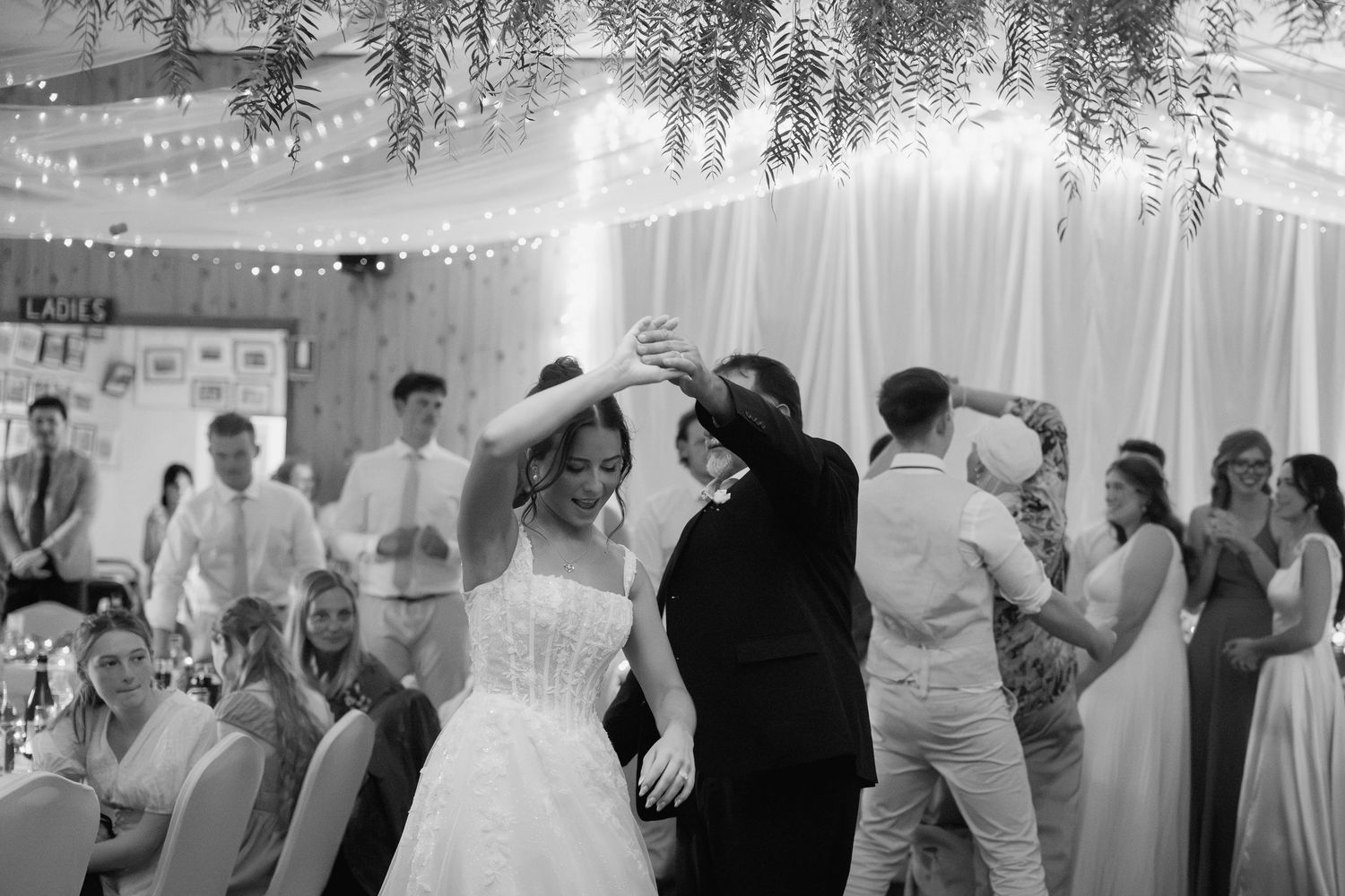 Black and white photo of bride and guests dancing joyfully at a wedding reception under fairy lights.