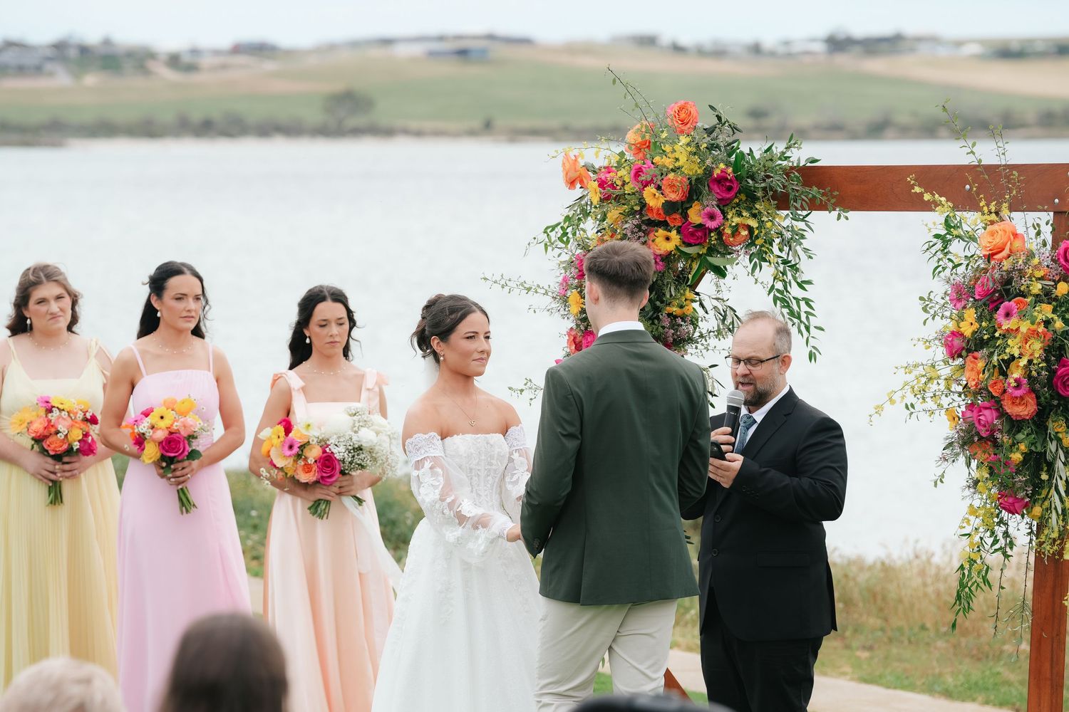 Outdoor wedding ceremony by a lake with bridesmaids in pastel dresses and a colorful floral arch.