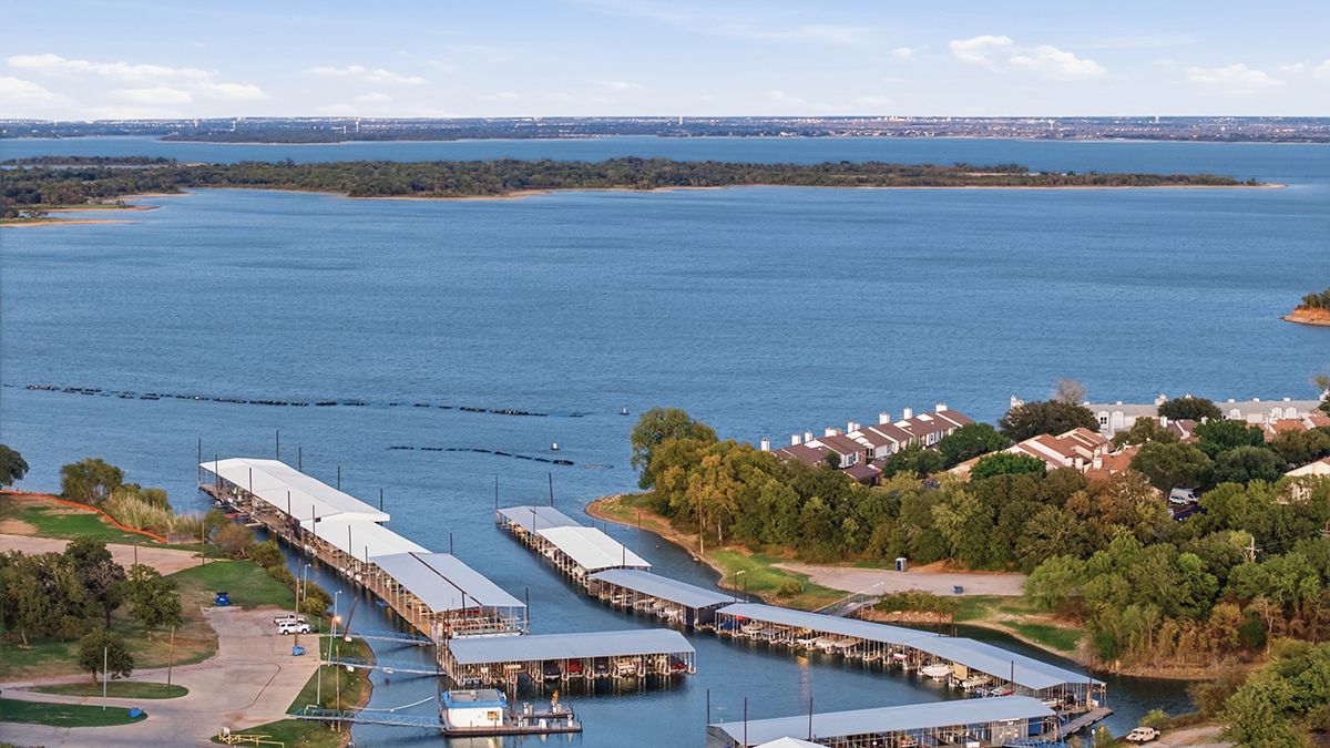 Aerial view of a covered marina with boat docks on a calm blue lake, surrounded by green trees, waterfront homes and a peninsula.