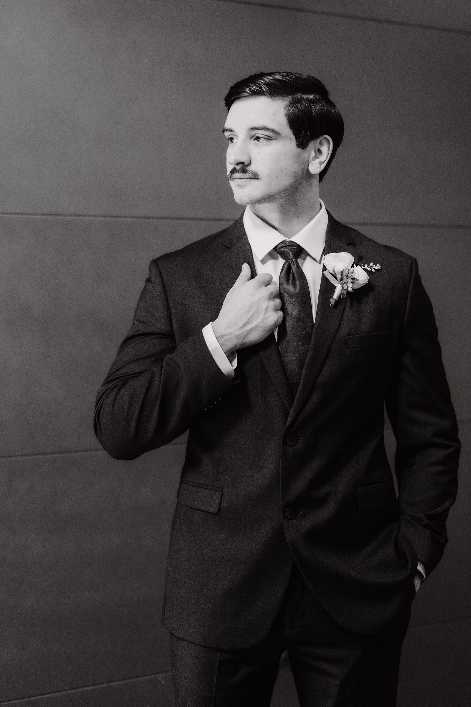 Black and white portrait of a groom in a dark suit with a floral boutonniere, adjusting his lapel and looking off to the side.