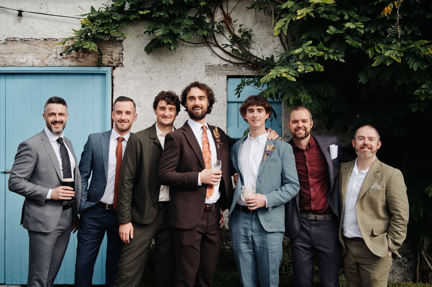 Seven groomsmen in suits posing together outdoors at a wedding, smiling in front of a blue door with ivy.