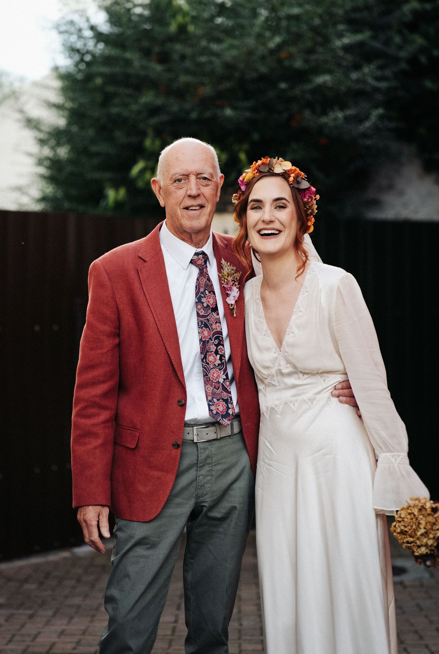 Bride in white dress and floral crown smiles with older man in red blazer and floral tie at outdoor wedding.