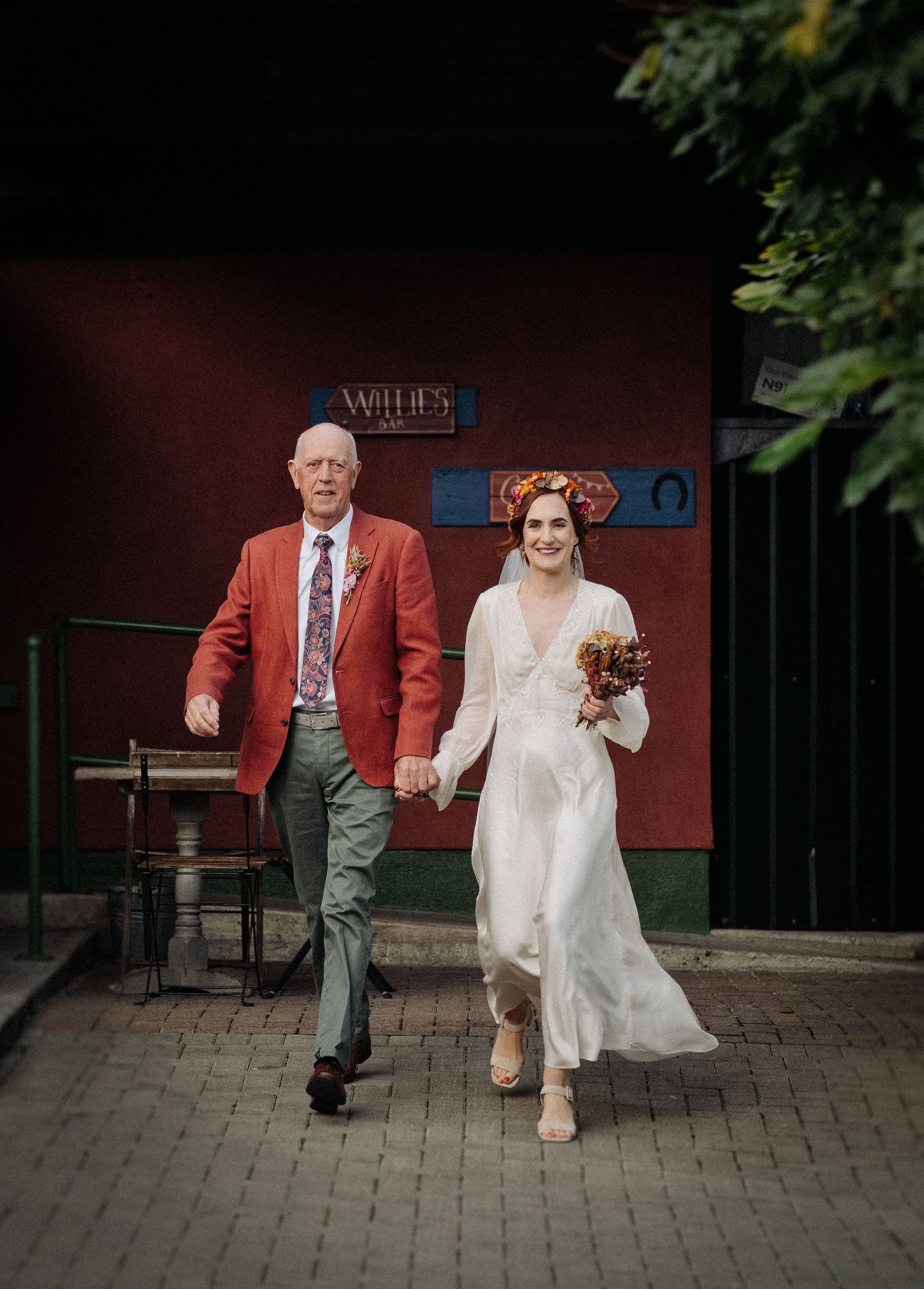 Bride in white gown with floral crown holds bouquet while walking with older man in red blazer outside rustic venue.