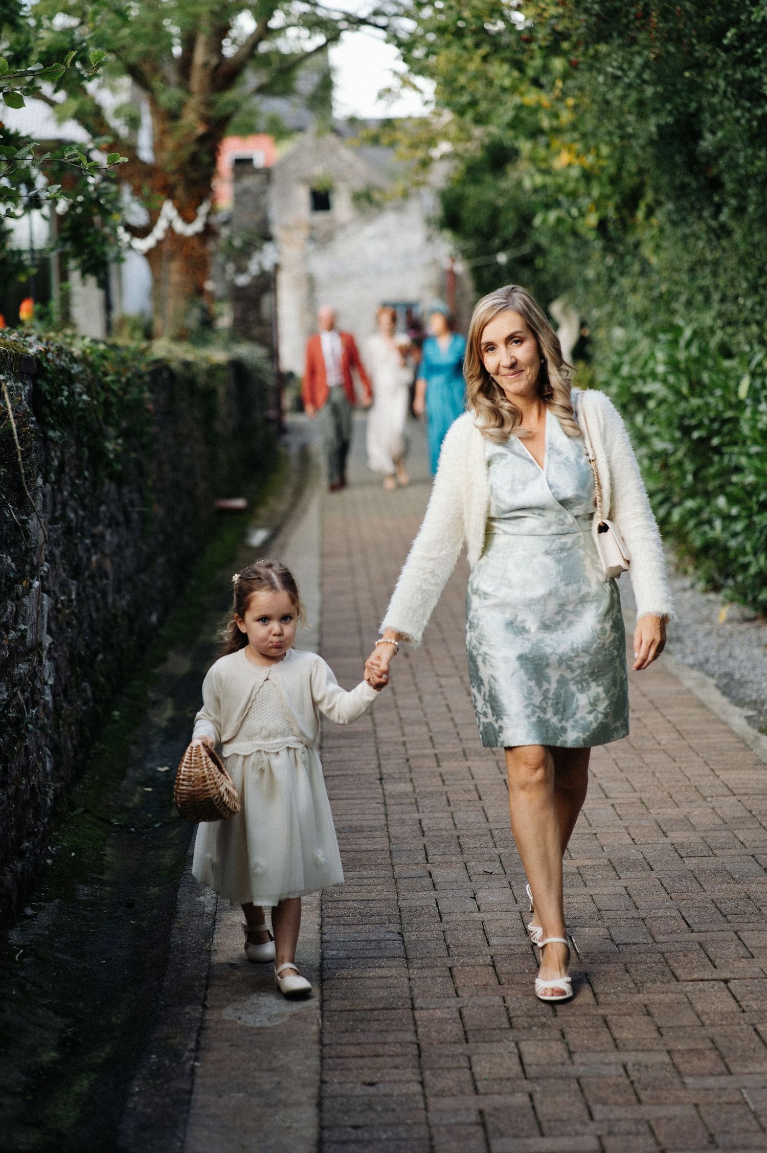Woman and young girl in elegant dresses walk hand-in-hand down a brick path at a wedding venue.