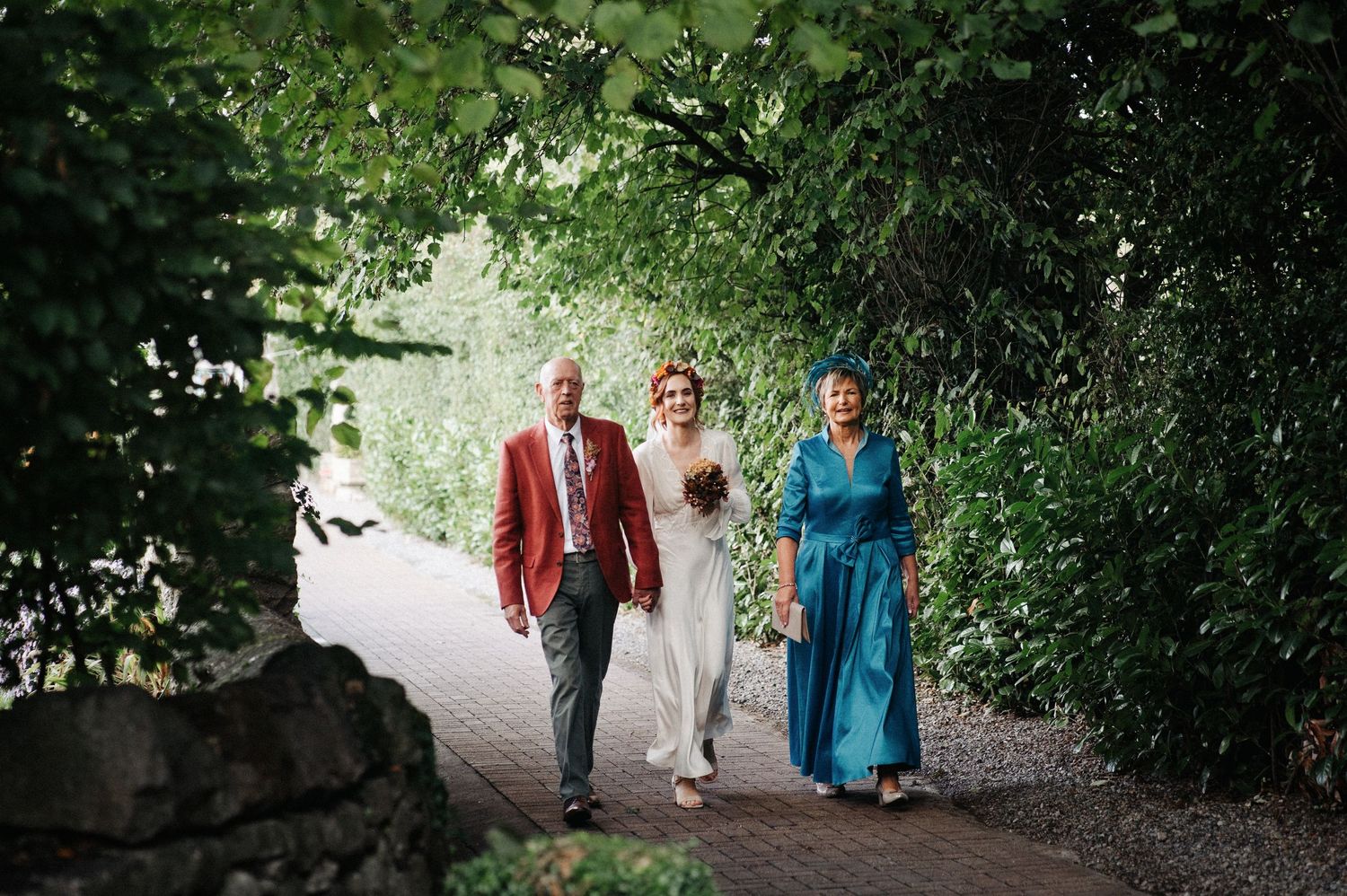 Bride in white dress walks with parents through lush green garden archway on wedding day.