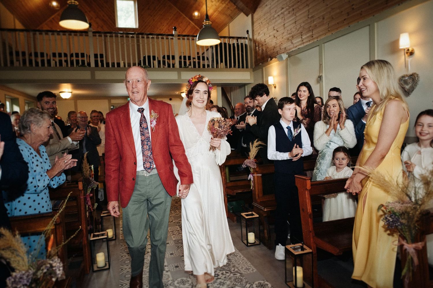 Bride walks down the aisle with older man in red jacket during wedding ceremony in rustic church.