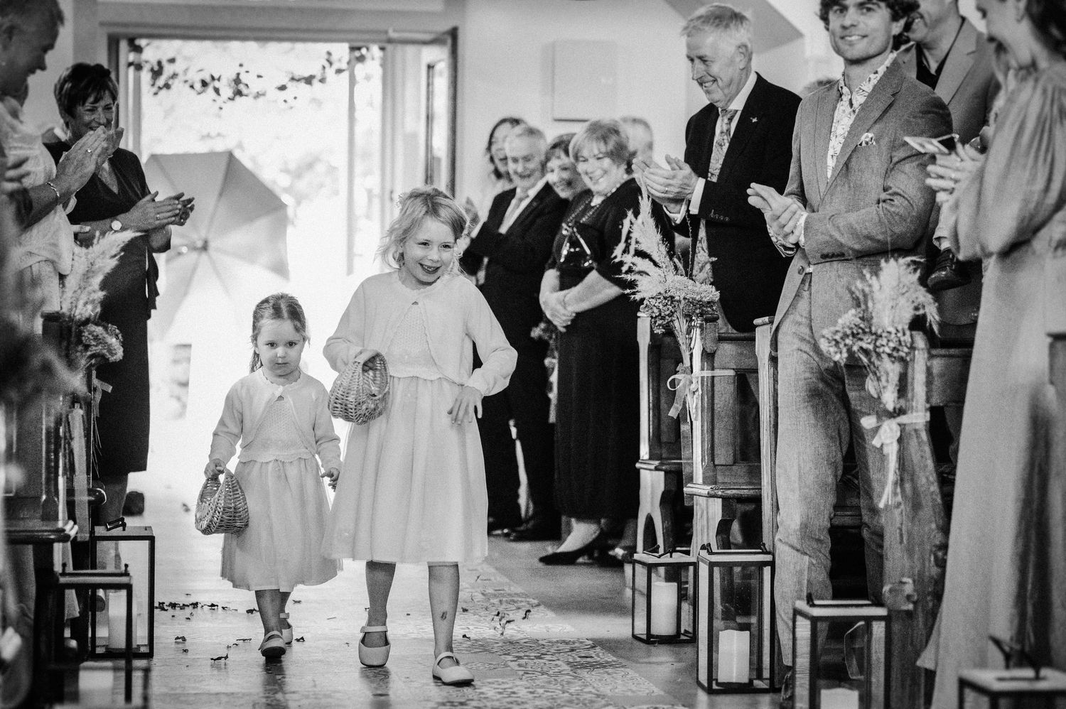 Two flower girls walk down the aisle at a wedding ceremony as guests applaud in black and white photo.