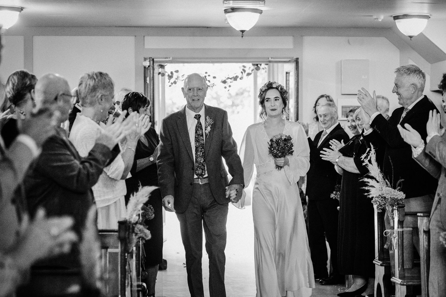 Bride walks down the aisle with her father at a wedding ceremony as guests applaud in black and white photo.