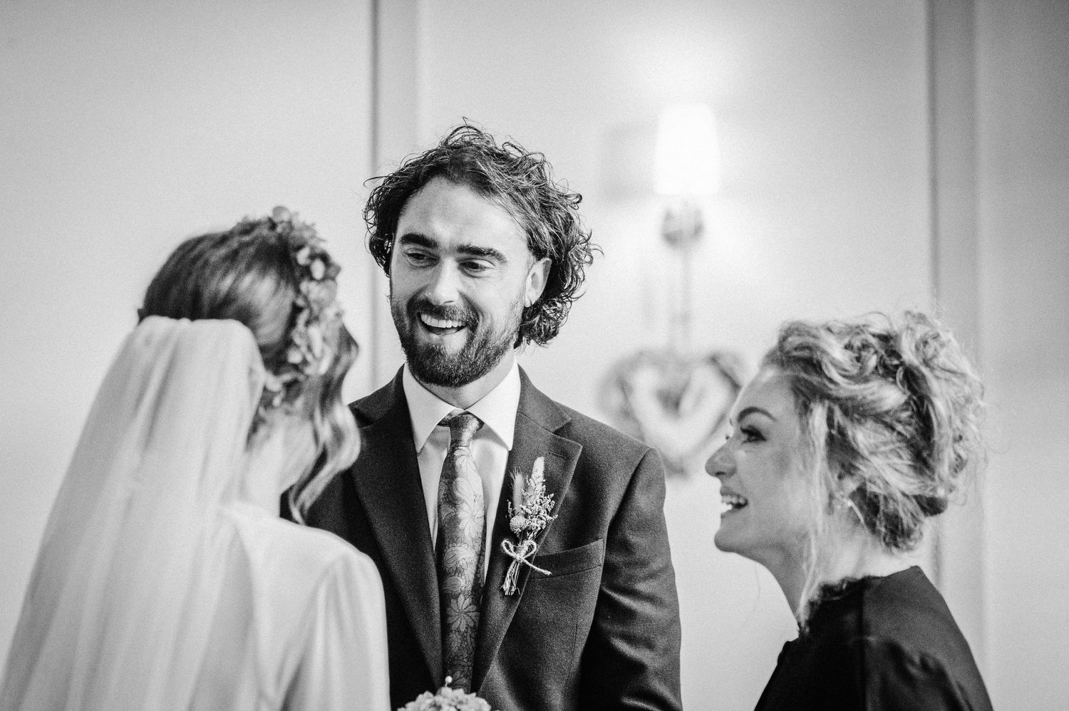 Black and white wedding photo of smiling groom in suit with boutonniere laughing with bride and guest.
