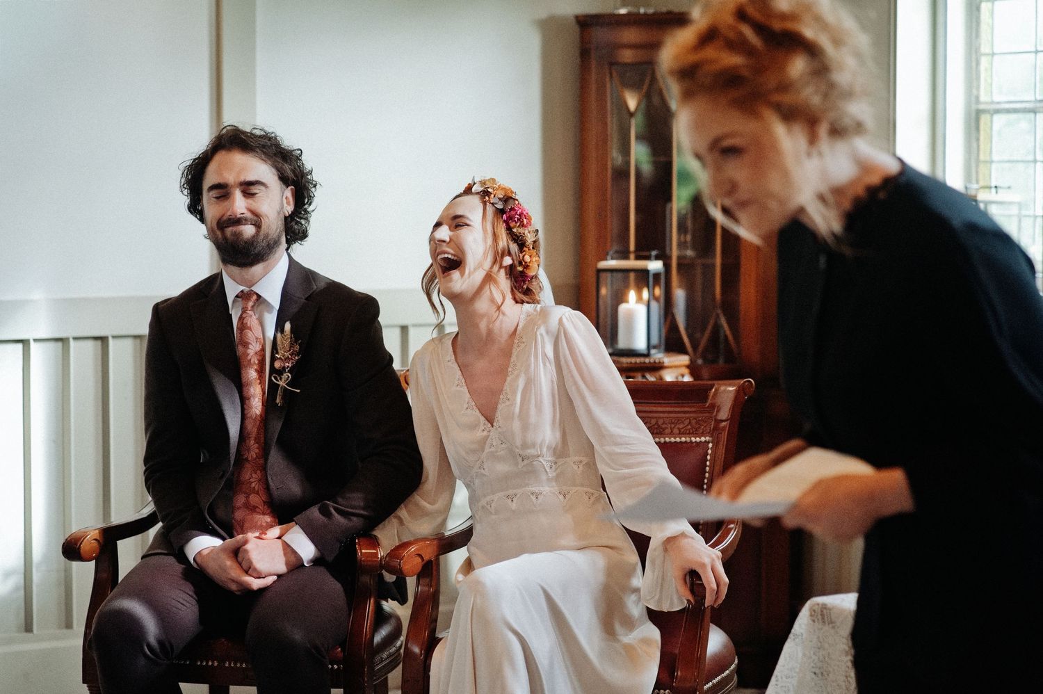 Bride laughing joyfully with groom during wedding ceremony, seated in elegant chairs in bright room.