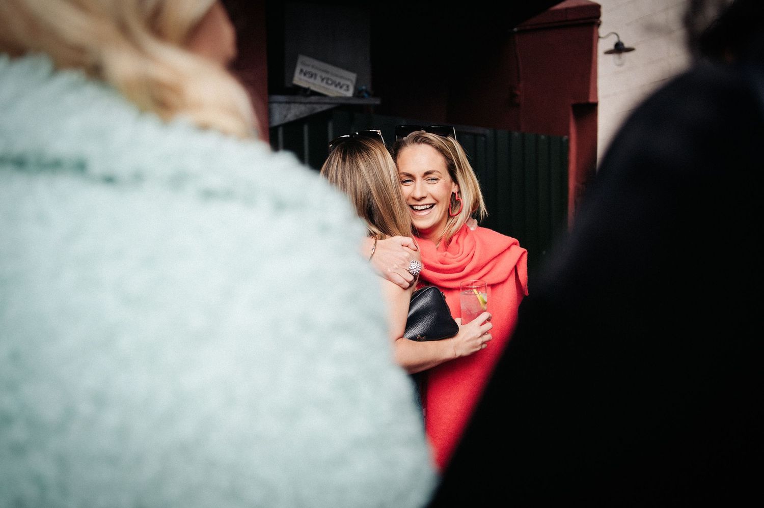 Two women sharing a warm hug and smiling at an indoor social gathering, one wearing a bright coral red top.