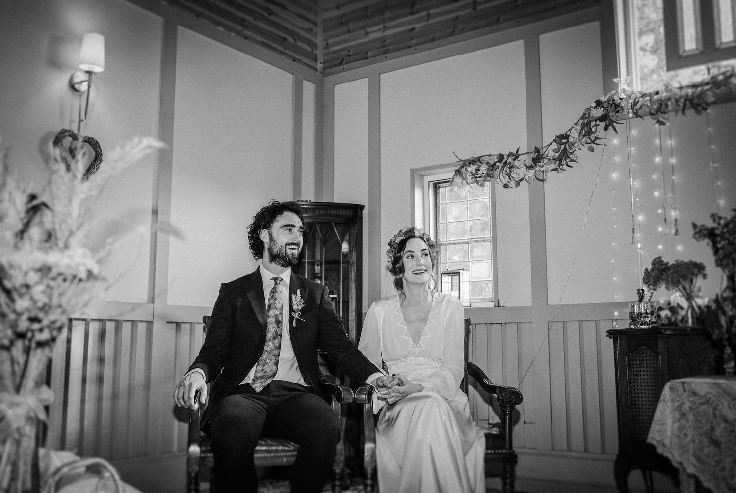 Black and white wedding photo of bride and groom seated together in a decorated room with string lights.