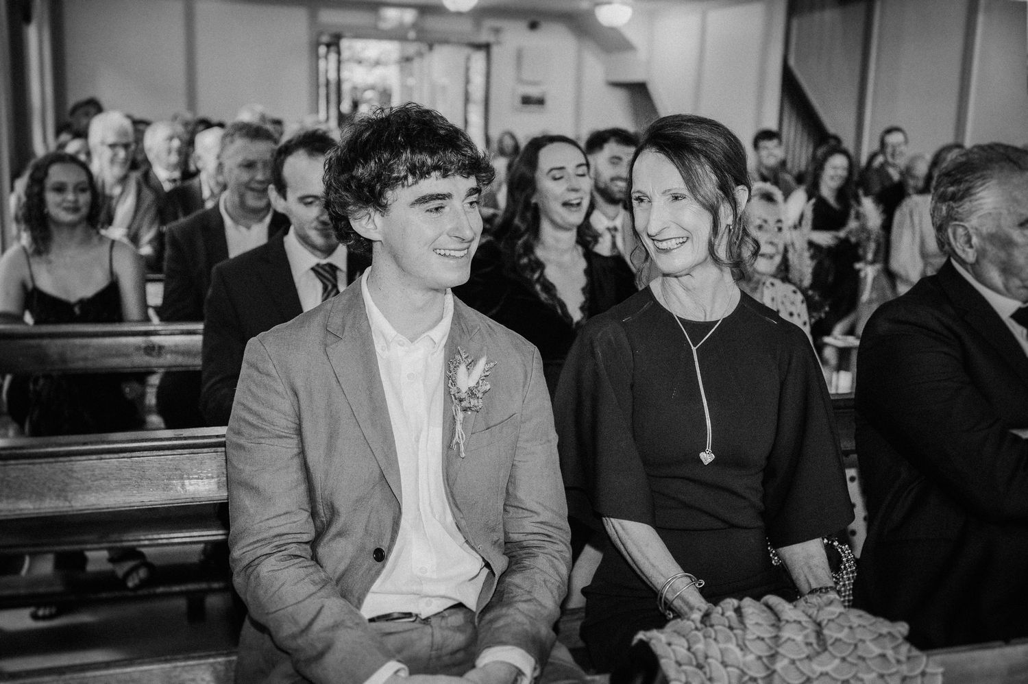 Young man in suit with boutonniere sits with smiling woman during wedding ceremony in black and white photo.