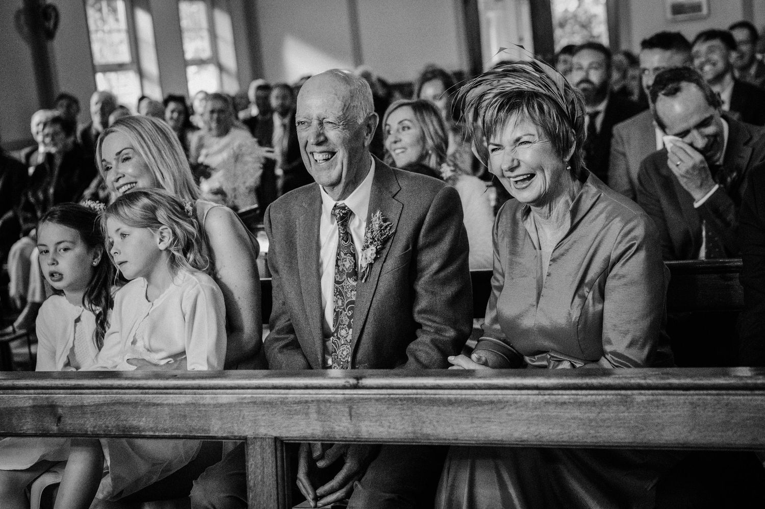 Black and white photo of smiling wedding guests seated in church pews during a ceremony.