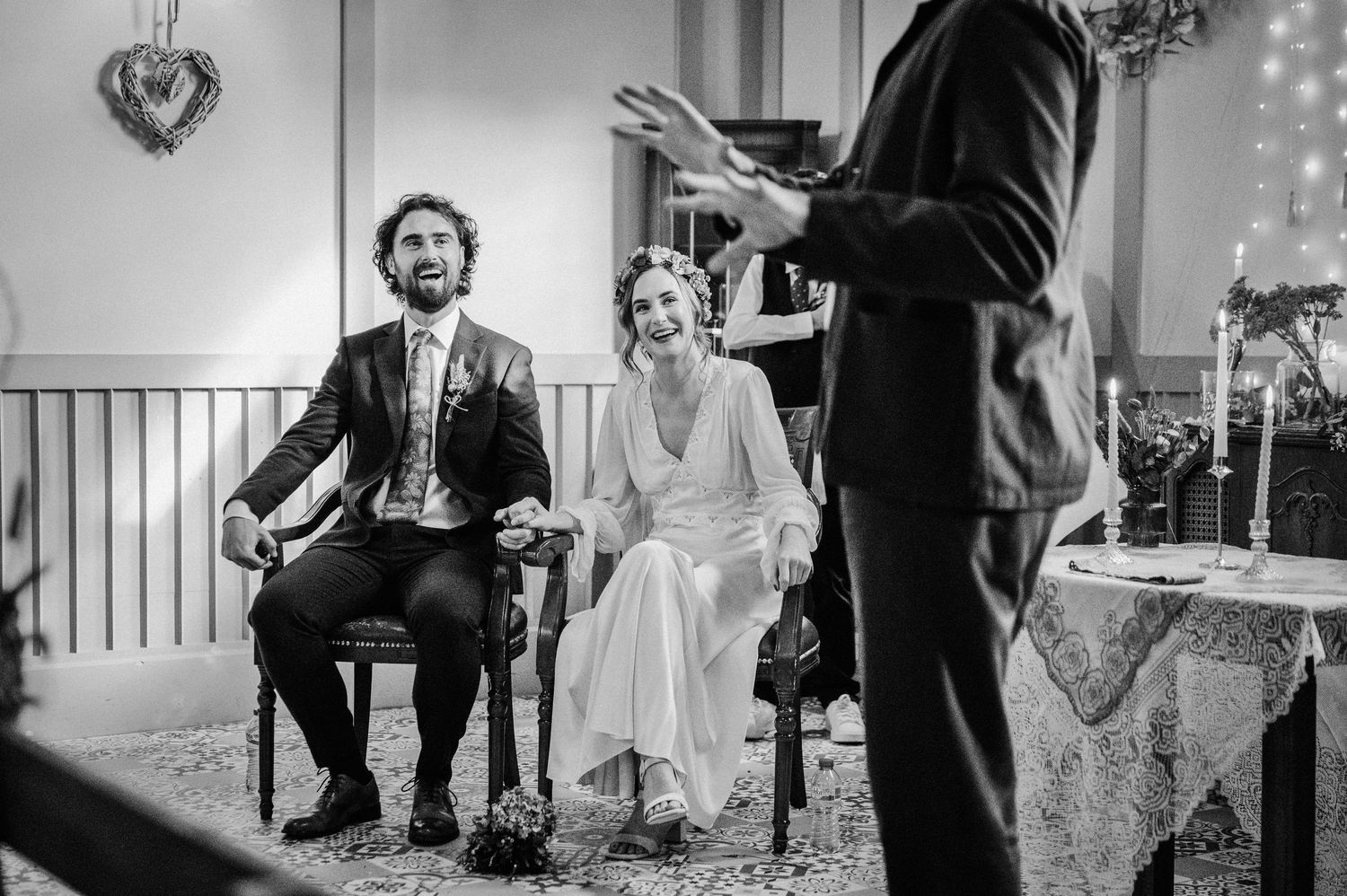 Black and white wedding photo of smiling bride and groom seated during ceremony while officiant speaks.