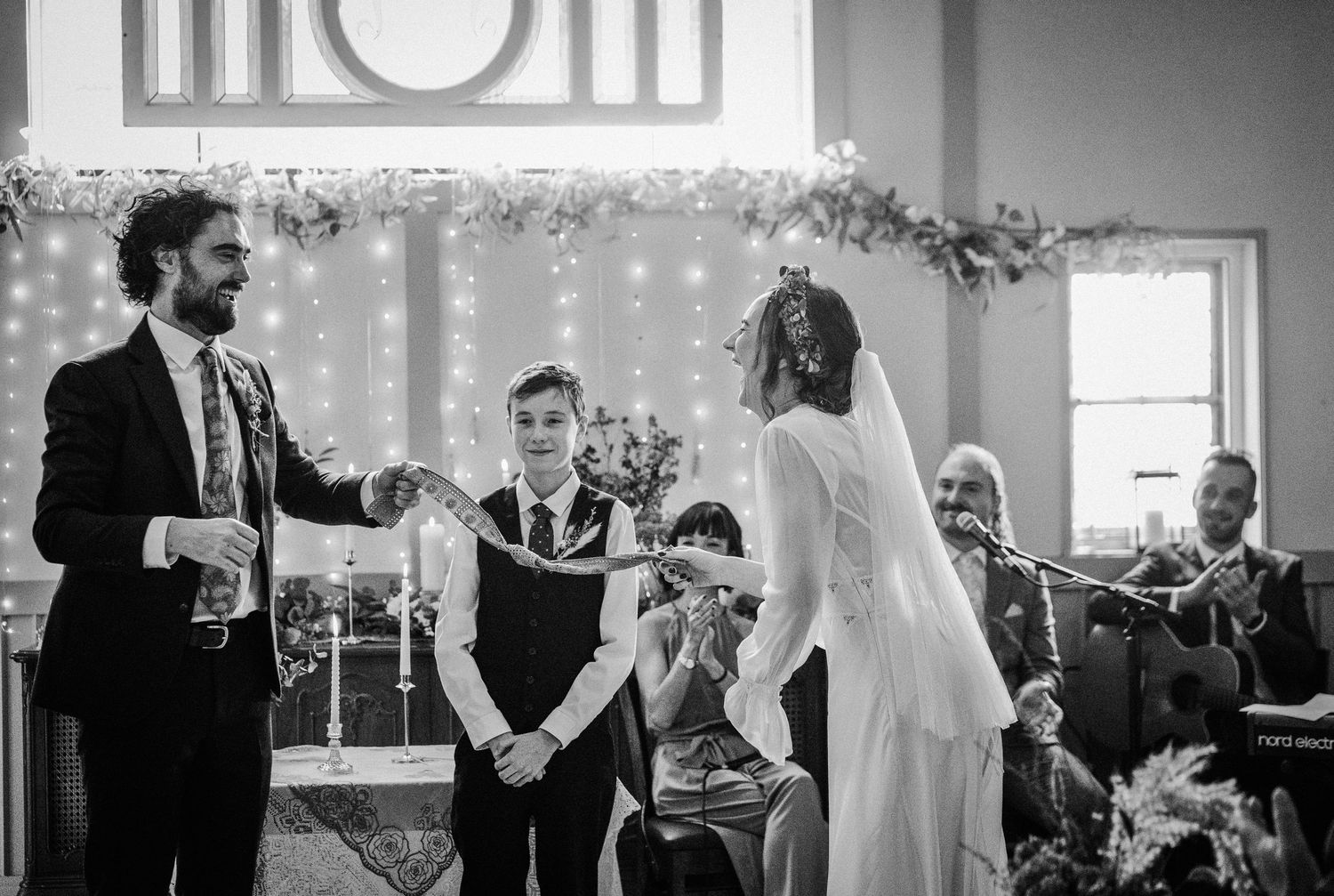 Black and white wedding ceremony photo of couple exchanging vows at altar with guests and officiant present.