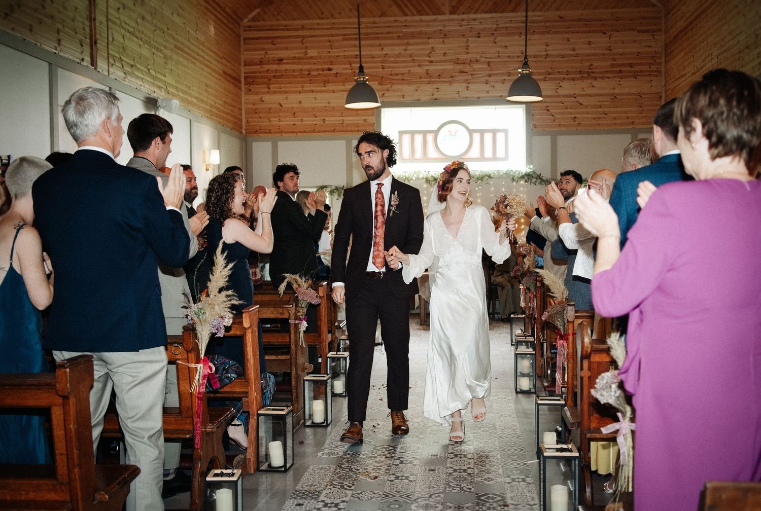 Joyful newlyweds walk down the aisle as guests applaud in a rustic wooden wedding venue.