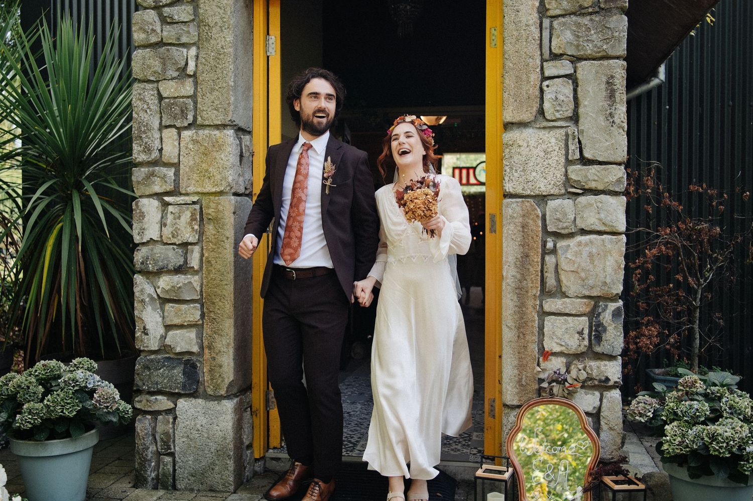 Happy newlywed couple holding hands outside a stone building with a yellow door on their wedding day.