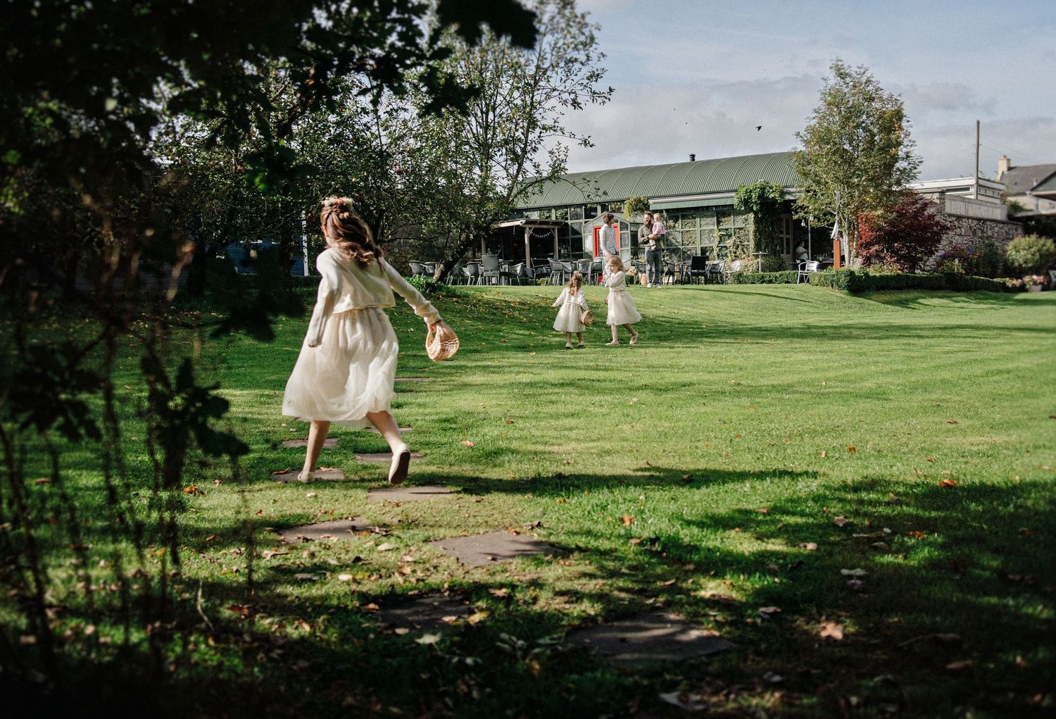 Flower girls in white dresses running across a green lawn at an outdoor wedding venue on a sunny day.