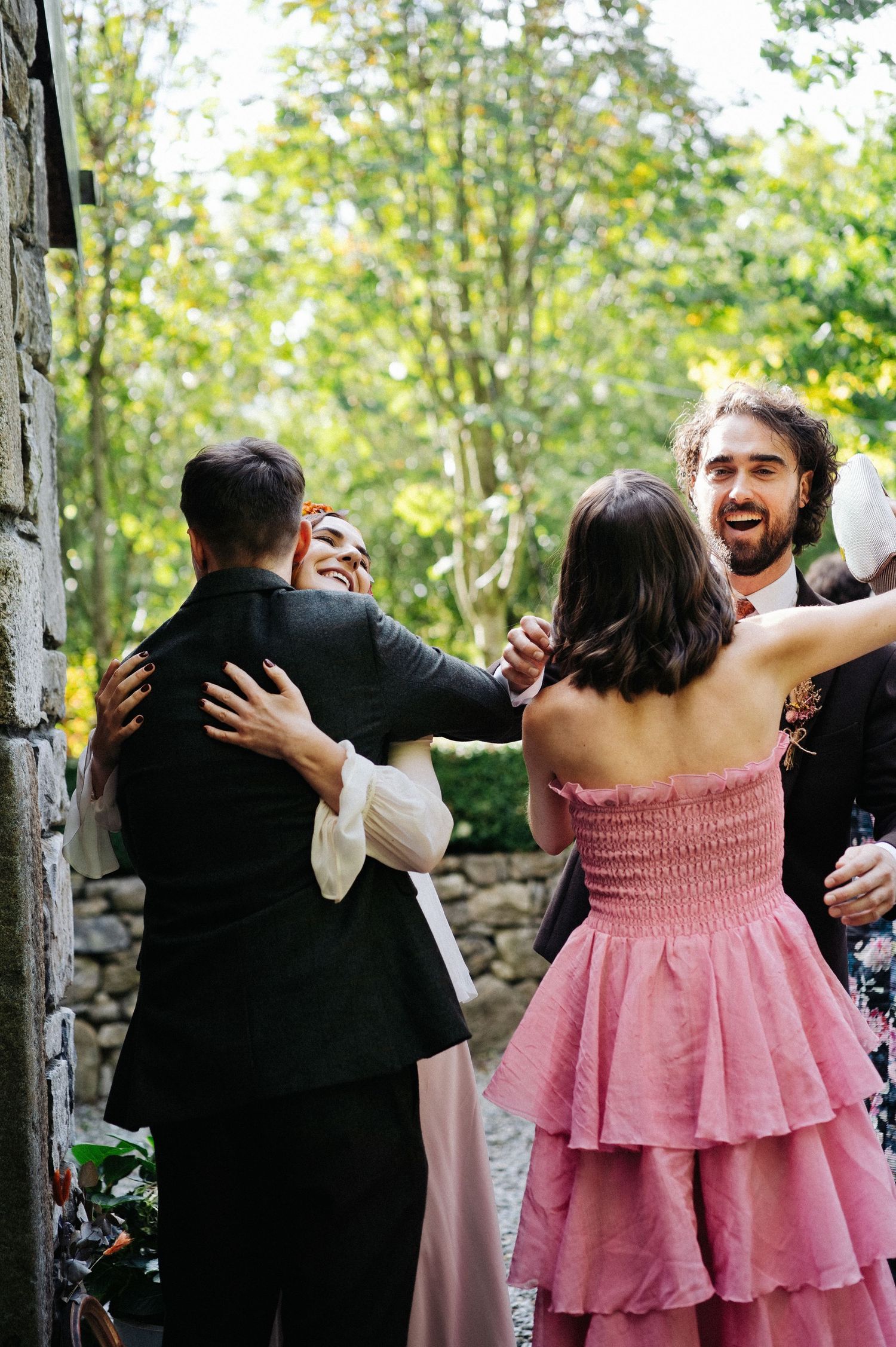 Guests in formal attire embrace and celebrate outdoors at a wedding reception near a stone wall.