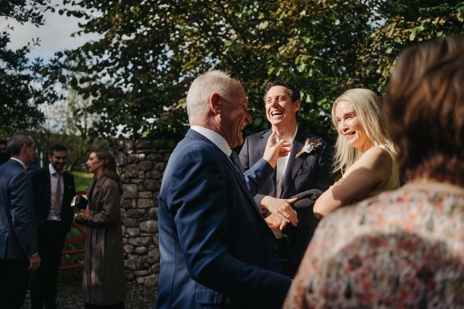 Guests laughing and socializing outdoors at a wedding reception, dressed in formal attire with lush green trees in background.