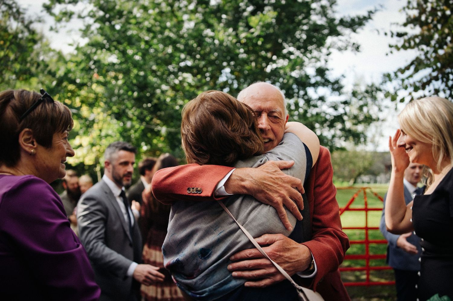 Elderly man in red jacket sharing warm embrace with guest at outdoor wedding reception among trees.