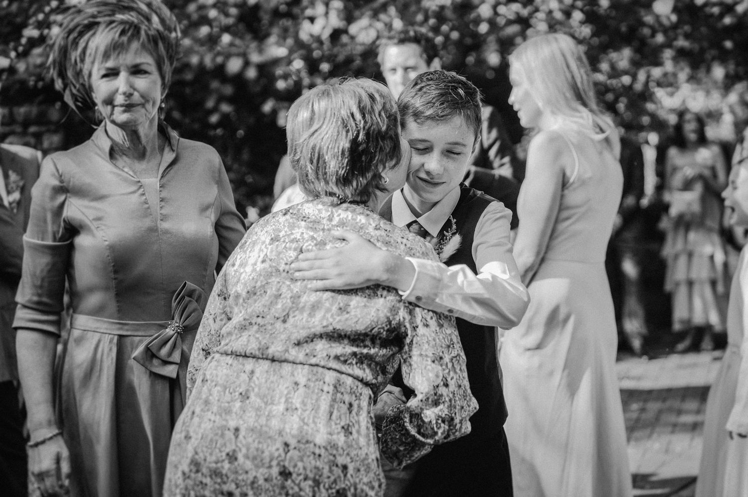A young groom embraces an elderly guest at an outdoor wedding ceremony while others look on warmly.