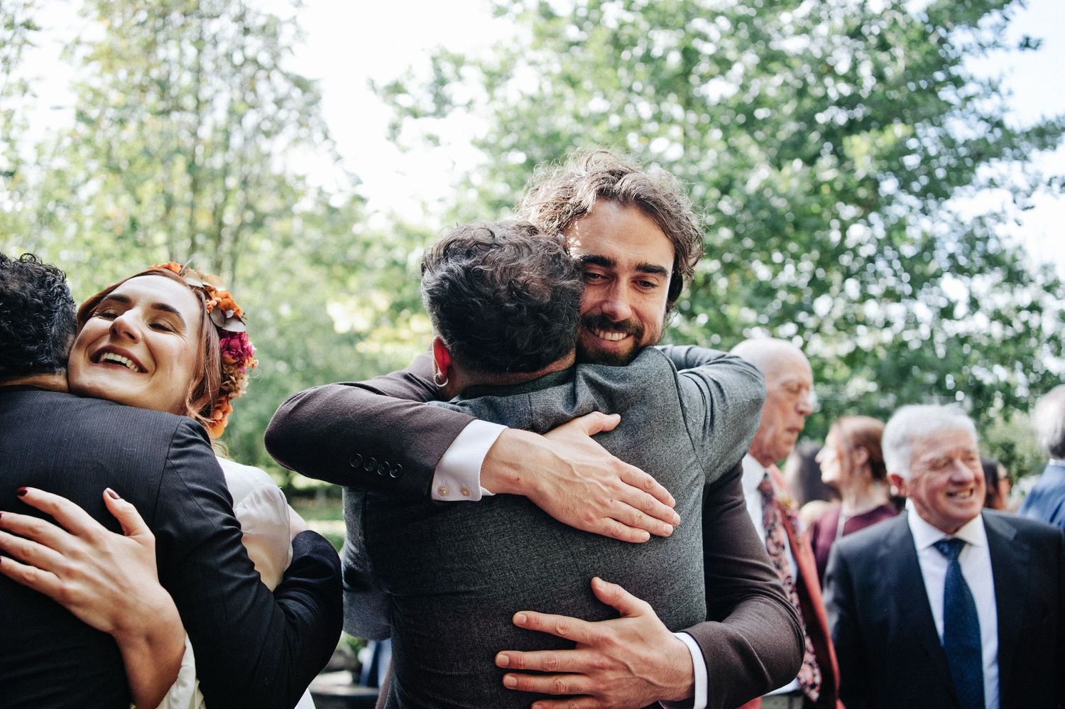 Guests sharing warm hugs and laughter at an outdoor wedding celebration surrounded by trees.