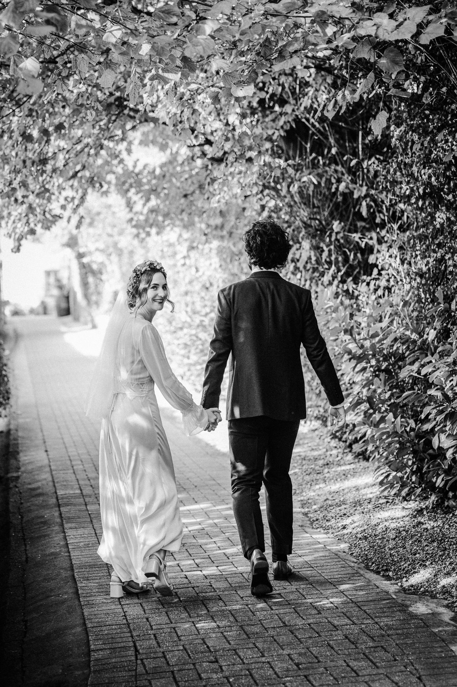 Black and white photo of a bride and groom walking hand in hand down a brick path lined with lush green foliage.