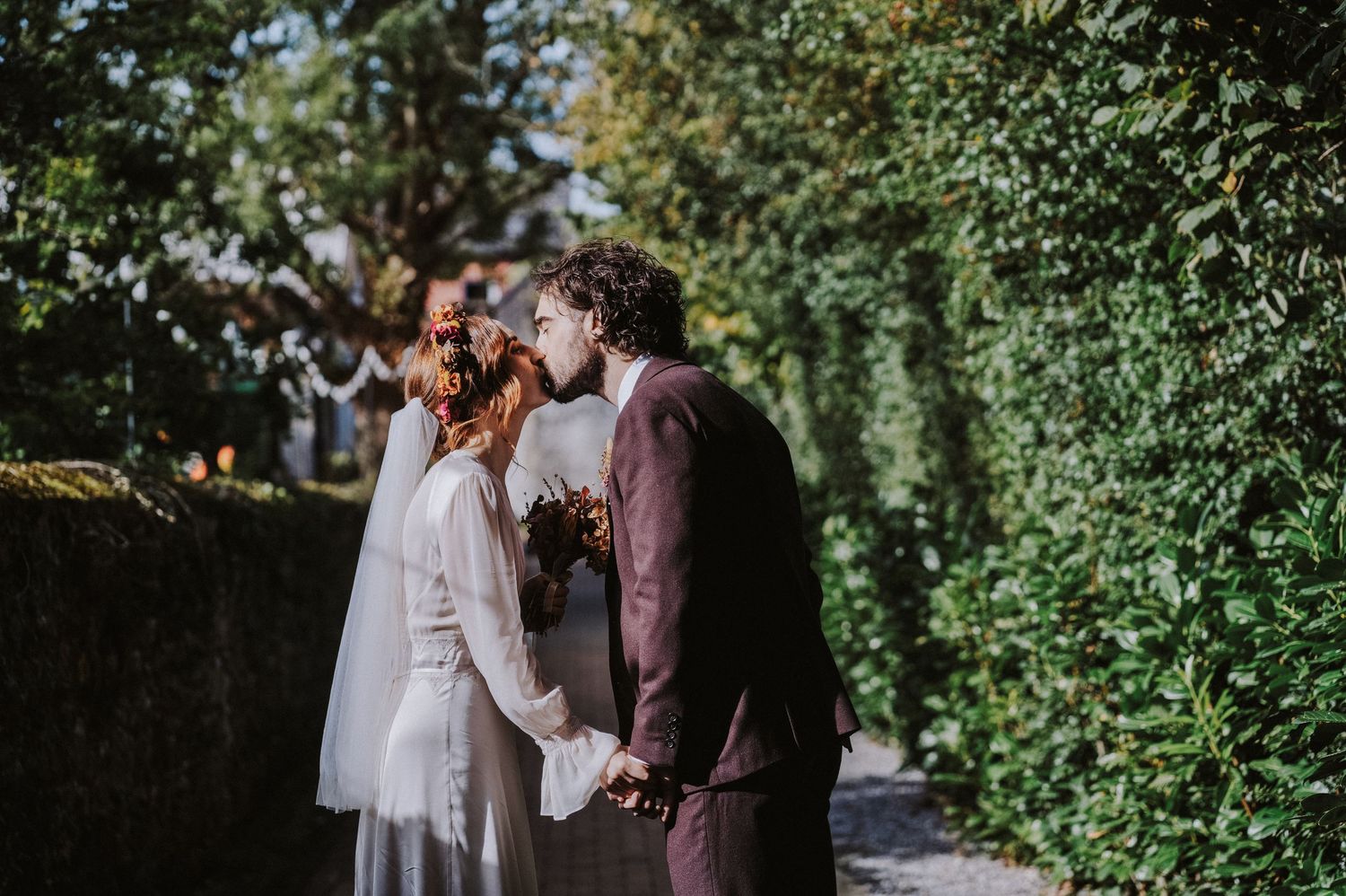 A bride and groom share a kiss while holding hands in a lush green garden on their wedding day.