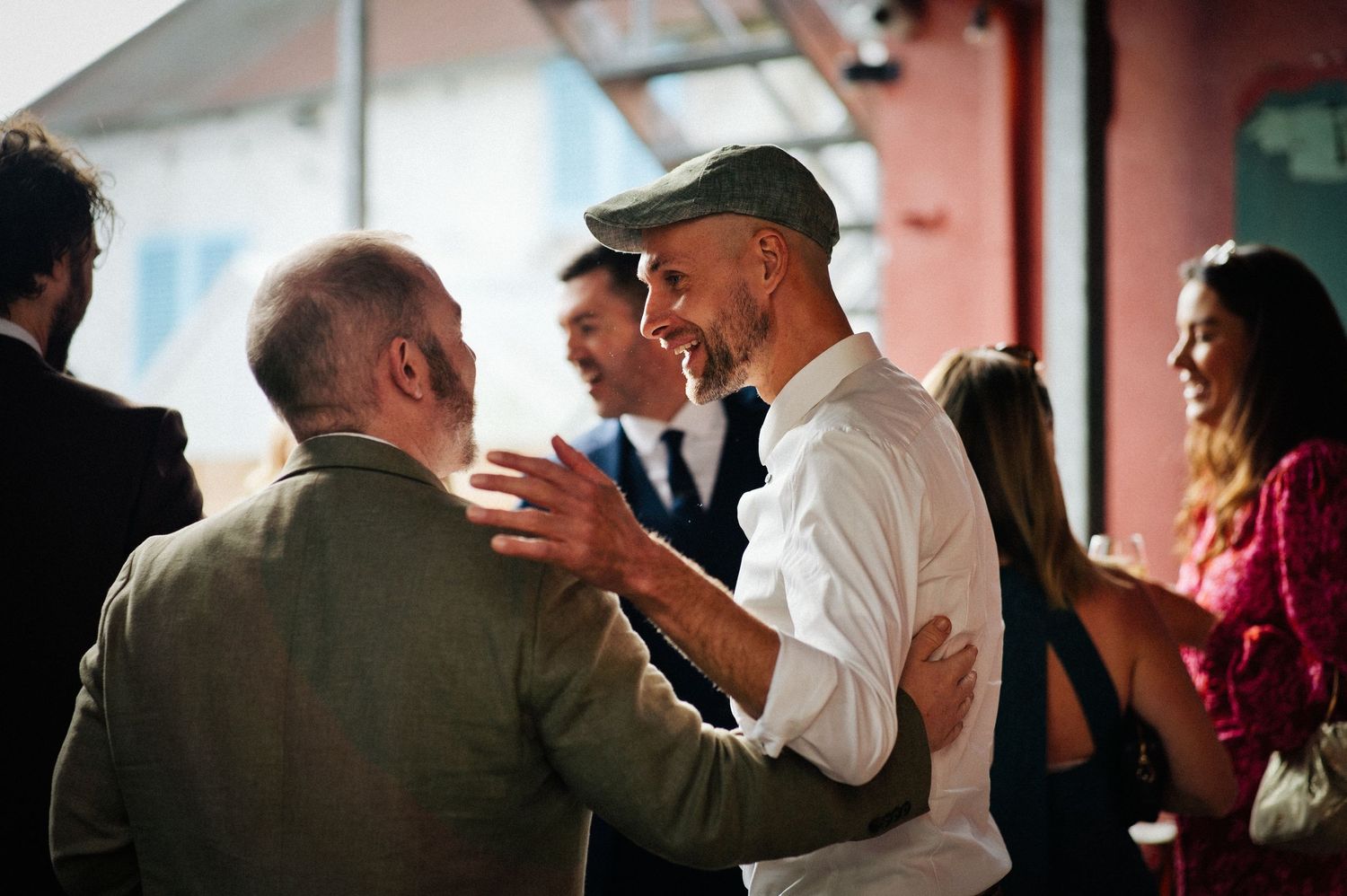 Two men greeting each other warmly at a social gathering, one wearing a green flat cap and white shirt.