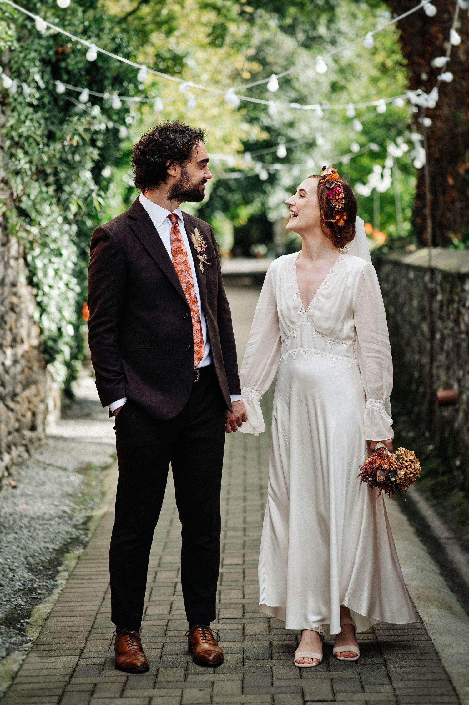 A bride and groom walk hand in hand on a cobblestone path, surrounded by lush greenery and string lights.